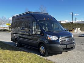 A black 2017 Ford Transit van with a large roof and chrome accents parked on a concrete surface with grass in the foreground