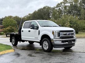A 2019 Ford F-350 Super Duty truck with a white cab and black flatbed parked at an angle with prominent wheels and chrome grille