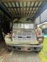 A 1985 Ford B600 truck with a green camouflage pattern and a distinctive front grille is parked under a shelter