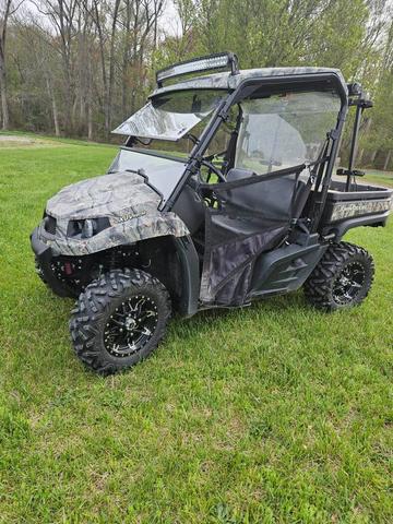 2015 John Deere 590m Gator in camouflage with a windshield and open door sitting on grass with large tires and a flatbed in the back