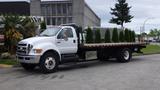 A white 2008 Ford F-750 flatbed truck with a row of small evergreen trees on the flatbed