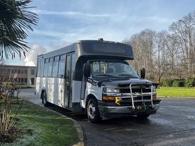 A 2018 Chevrolet Express bus with a white exterior and black bumpers is parked, featuring a grille guard and large windows on the side