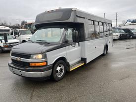 A 2017 Chevrolet Express bus with a gray and white exterior and large windows parked on a wet surface