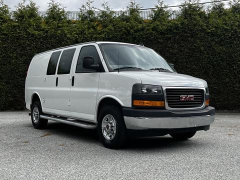 A 2023 GMC Savana van in white with a distinctive grille featuring the GMC logo and chrome accents, equipped with four doors and a silver wheel trim