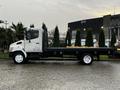 A white 2006 Hino 155 flatbed truck with a black bed parked on a wet surface with trees in the background