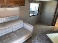 A beige tufted couch is positioned near a window in a camper, with a textured gray wall and a light-colored carpet visible in the foreground