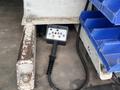 A control panel with various buttons and a handle is resting on the floor of a vehicle, next to a rusted metal beam and blue storage bins