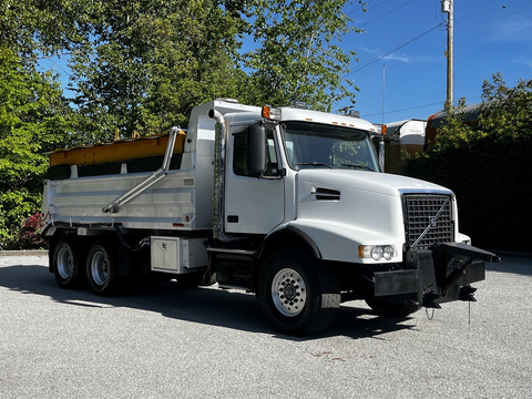 A 2005 Volvo VND 122 dump truck with a white cab and silver cargo bed parked with a raised dump feature and front plow attachment