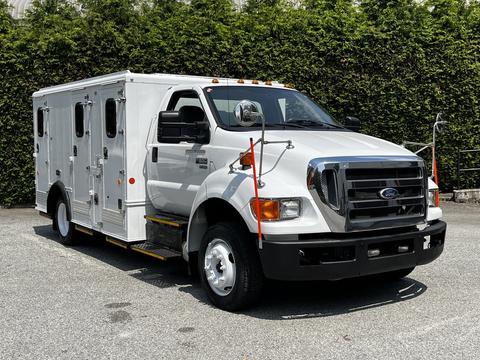 A 2011 Ford F-650 truck with a white body and multiple side doors is parked with a chrome front grille and orange lights on the roof