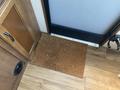 A rectangular doormat made of coir placed in front of a door inside a camper with wooden flooring and cabinetry nearby