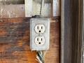 A weathered electrical outlet mounted on a wooden wall with exposed wiring hanging below it