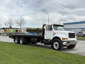 A white 2000 International 4900 flatbed truck with a black flatbed and three axles parked on a grassy area