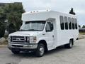 A white 2014 Ford E-350 bus with large windows and a boxy design is parked in an outdoor setting