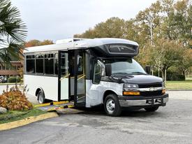 A 2019 Chevrolet Express shuttle bus with a black and white exterior features a wheelchair ramp deployed at the entrance and has multiple windows on the side