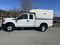 A white 2016 Ford F-250 Super Duty truck with a camper shell on the bed parked on a gravel surface