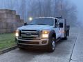 A white 2015 Ford F-550 with a flatbed and orange marker lights is parked on a street with a foggy backdrop