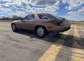 A 1983 Porsche 928 in a metallic bronze color parked on a road with distinctive round headlights and a sleek design showcasing its sporty profile and unique rear styling