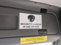 A sign displaying a security camera with the text "SECURITY CAMERA IN USE ON BUS" mounted on the interior of a bus
