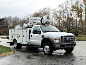 A white 2008 Ford F-550 with a bucket truck mounted on top and a utility box on the side is parked on a wet surface