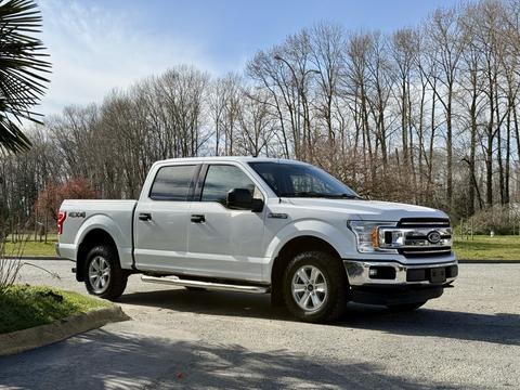 A white 2018 Ford F-150 truck with chrome accents and four doors parked on a gravel driveway