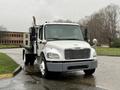 A 2019 Freightliner M2 truck with a flatbed, featuring a single exhaust pipe and multiple lights on the front, is parked on a wet surface.