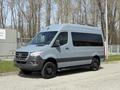 A 2019 Mercedes-Benz Sprinter van with a gray exterior and tinted windows parked alongside a road with trees in the background