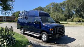 A blue 2013 Ford Econoline van with a high roof and a ladder rack on top parked on a driveway