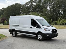 A 2022 Ford Transit van in white with a black stripe on the lower side parked at an angle with its sliding door closed and alloy wheels visible