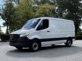 A 2020 Mercedes-Benz Sprinter van with a white exterior and black accents parked in a lot with trees in the background
