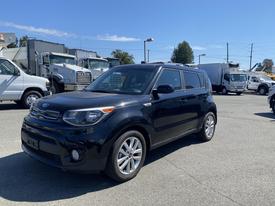 A black 2017 Kia Soul parked at an outdoor location with a clear sky and several large vehicles in the background