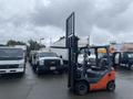 A 2011 Toyota 028FG15 forklift with an upright fork assembly is positioned in a parking area among several trucks and other vehicles