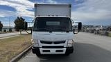 A white 2018 Mitsubishi Fuso FE box truck with an enclosed cargo area and orange roof lights parked on a road