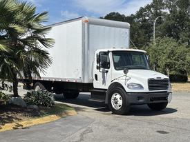 A white 2015 Freightliner M2 106 box truck is positioned in the foreground with a silver cargo area and visible headlights and grill