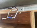 A metal tool handle with a blue grip attached to a silver pole, mounted on a wooden shelf with a partially visible, plain wooden structure behind it