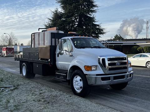 A white 2011 Ford F-750 truck with a flatbed and a brown tank on the back is parked with its headlights on and fog on the road