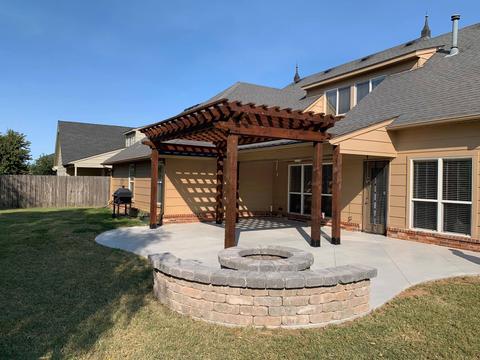 An outdoor fireplace made of stone with a circular design sits on a paved area under a wooden pergola providing shade and structure