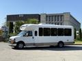 A 2017 Chevrolet Express bus with a white exterior and large windows parked on a street with palm trees nearby
