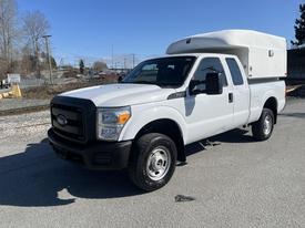 A 2016 Ford F-250 SD with a white cab and a camper shell on the bed parked on a gravel surface