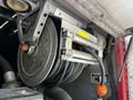 A metal hose reel with a circular drum and coiled black hose attached, mounted on a fire truck with visible metal and plastic components