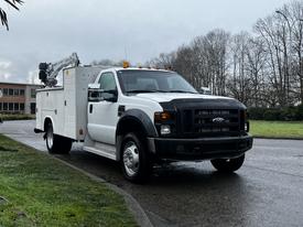 A 2009 Ford F-550 utility truck in white with a black front grille and a service body featuring a ladder rack on top and illuminated warning lights on the roof