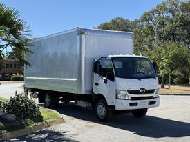 A white 2018 Hino 195 box truck parked on a street with a large cargo area and a cab with headlights on