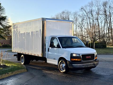 A white 2015 GMC Savana box truck with a flatbed and a large cargo area is parked at an angle with its headlights on