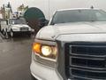 A white 2015 GMC Sierra 1500 pickup truck with a prominent front grille and one headlight illuminated