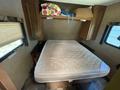 A double bed with a tan mattress is positioned in a camper interior with overhead storage and a small table on the side
