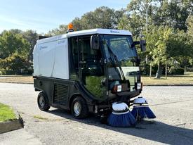 A 2017 Madvac LS100 street sweeper with dual front brushes and an orange flashing light on top parked on a street