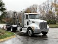 A 2020 Peterbilt 330 truck with a white cab and black flatbed attachment parked with warning lights on