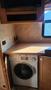 A compact washing machine is installed under a countertop in a recreational vehicle with wooden cabinetry and electrical outlets visible
