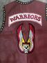 A maroon leather vest with the word "WARRIORS" prominently displayed at the top and an embroidered design featuring a skull surrounded by red and yellow wings at the center