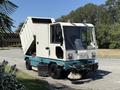 A 2001 Tennant 830-II street sweeper with a cab and large debris hopper raised at the back is positioned in the foreground with a broom attached at the front