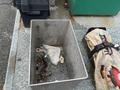 A metal container with some tools and a dirty cloth inside is placed on a textured surface next to a rolled canvas bag